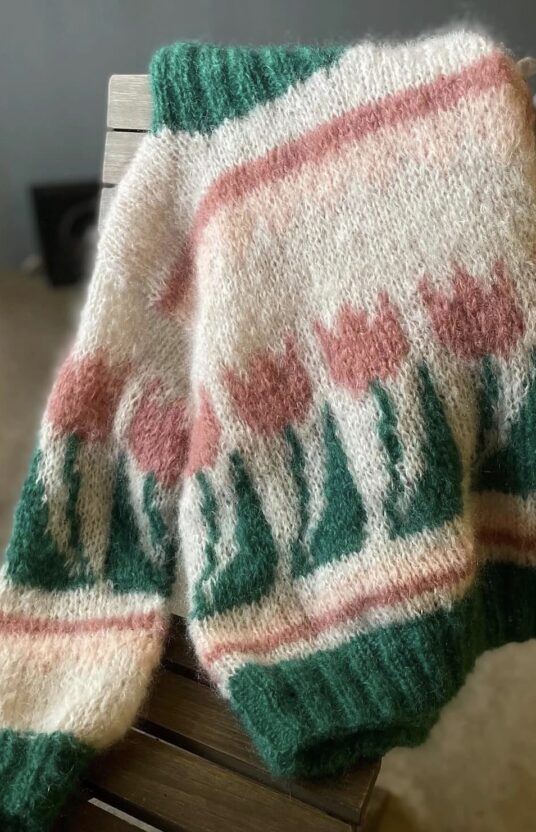 Colorful knit sweater with green cuffs and hem draped over a chair, featuring pink floral patterns on a cream background.