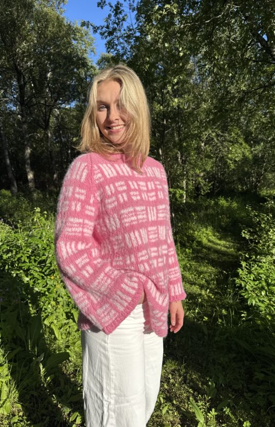 Smiling woman with blond hair in a pink fuzzy sweater and white pants, standing in a sunlit forest.
