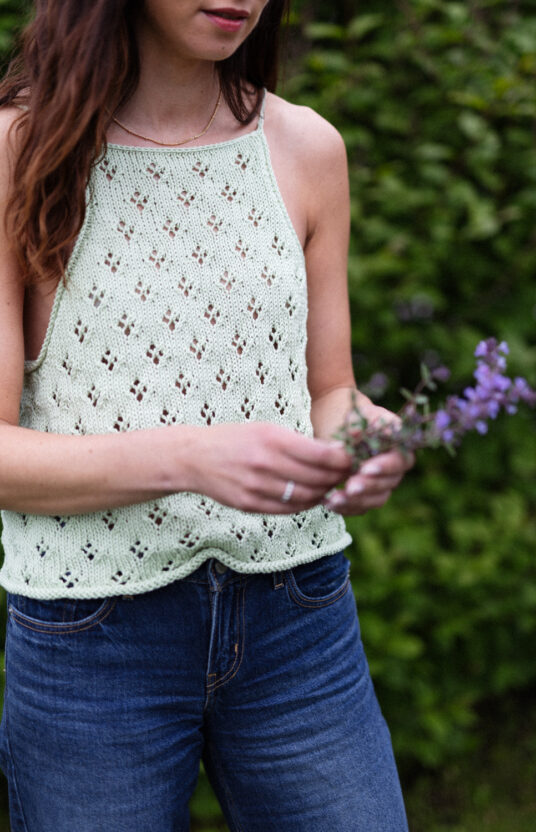 Woman in a mint crochet halter top and jeans, standing outdoors while holding sprigs of lavender in her hands.