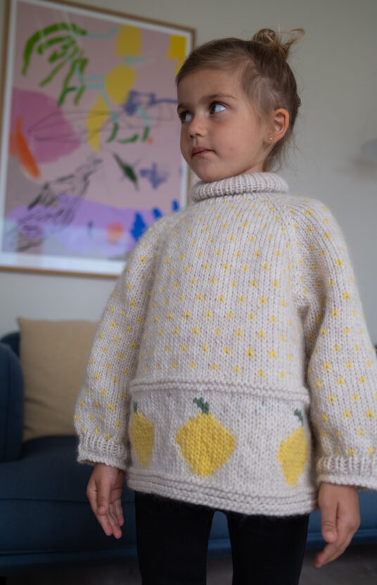 Young girl wearing a cream knitted sweater with yellow lemon shapes stands in a living room with abstract colorful artwork on the wall behind her.